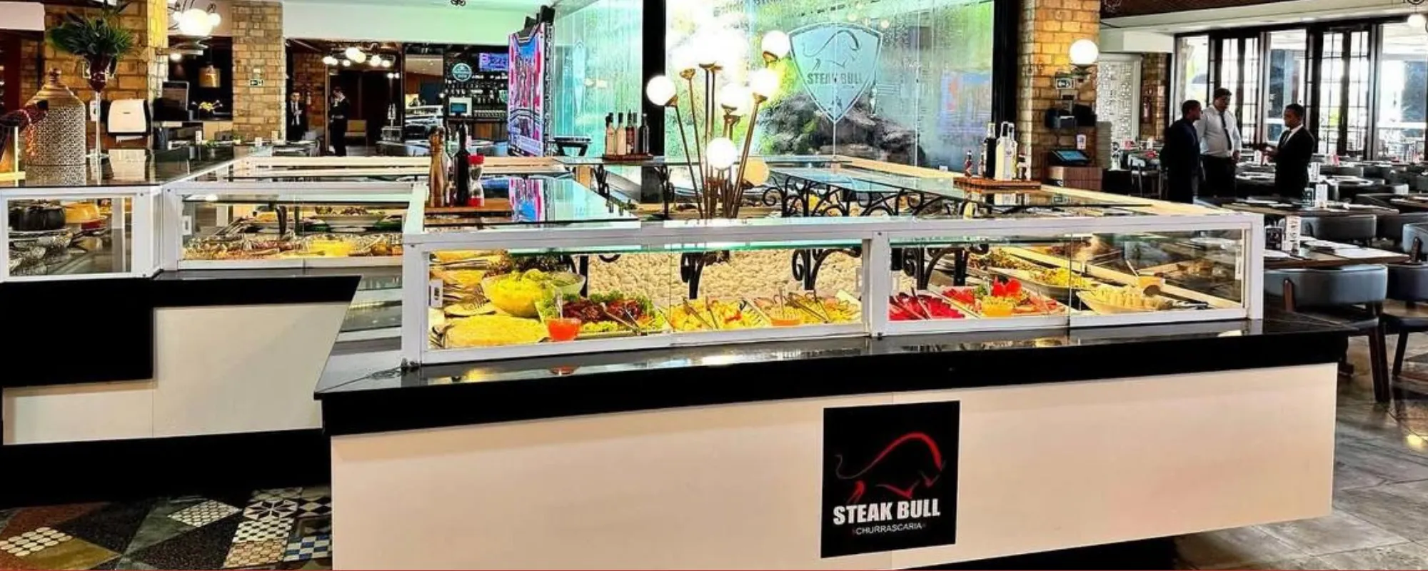 Salão do Steak Bull com o buffet refrigerado em primeiro plano e mesas ao fundo.